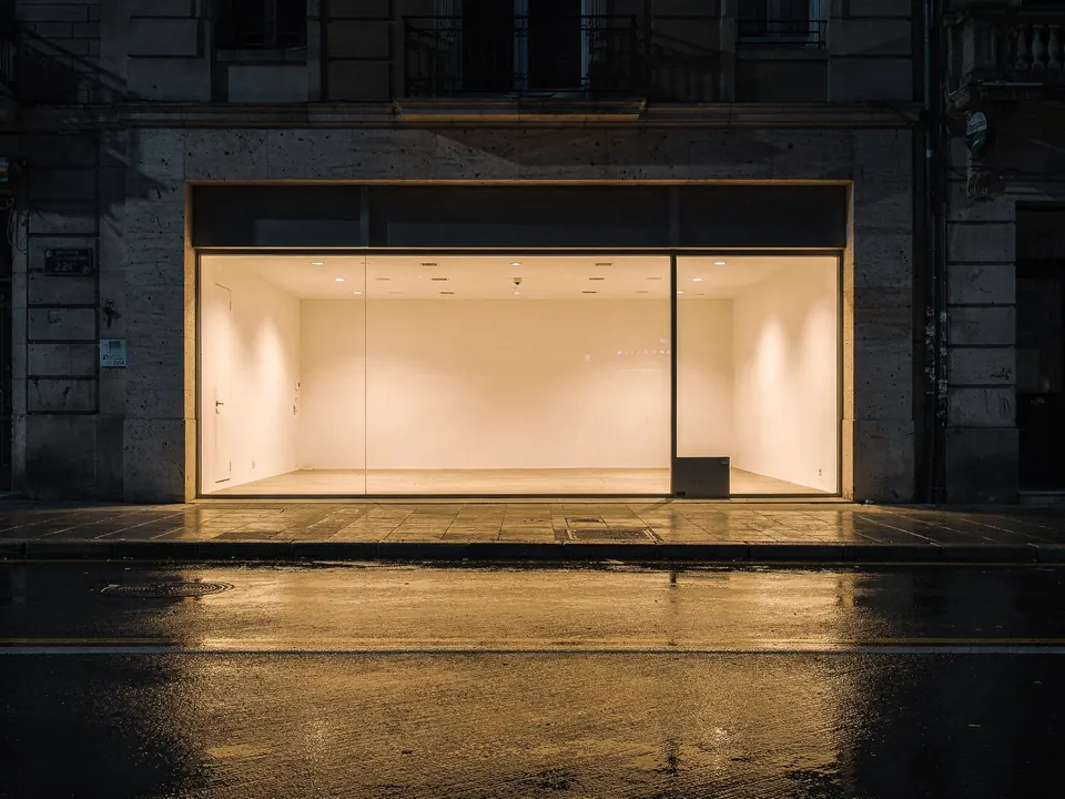 Local comercial vacío con escaparate de vidrio iluminado de noche, junto a una calle mojada